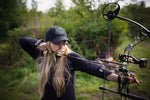 black hat with green and black precision shooter patch on blonde woman shooting bow